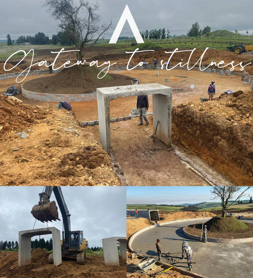 Entrance roundabout construction with circular landscaped island and concrete gateway arches, workers and excavator on-site