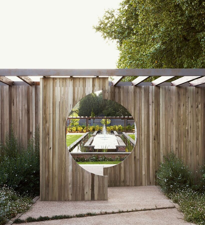 Private hotel courtyard with wooden pergola and circular cutout framing a long reflecting pool and fountain amid lush gardens.