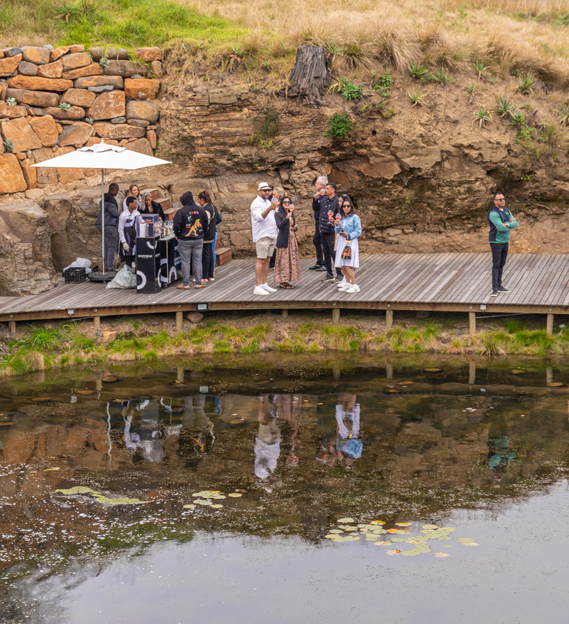 Waterside wooden terrace with bar and umbrellas; guests sip cocktails beside a rocky stone wall, relaxed alfresco by the pond.