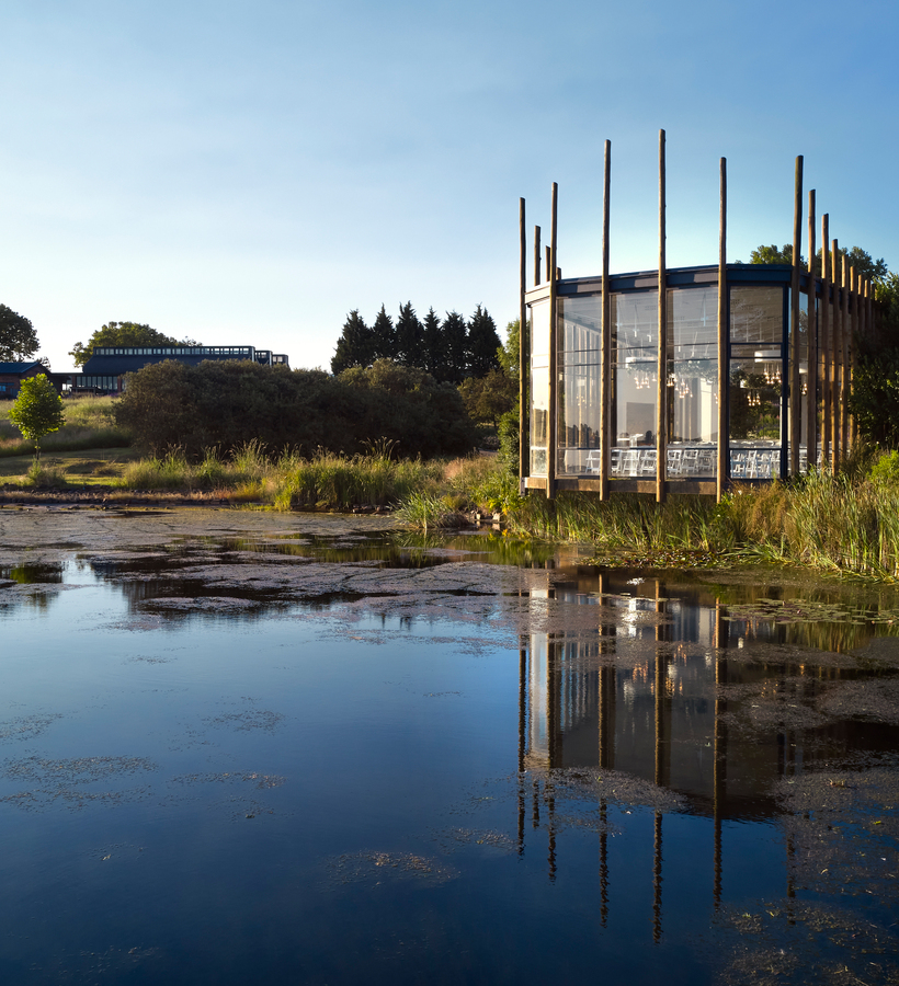 Lakeside glass dining pavilion on wooden stilts with chandeliers and white chairs, reflected in a calm pond amid grassy meadow.