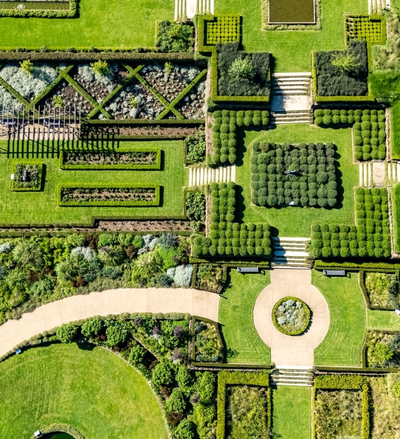 Aerial view of manicured formal gardens with geometric boxwood parterres, raised planting beds, stone steps and curved paved paths for guest strolls.