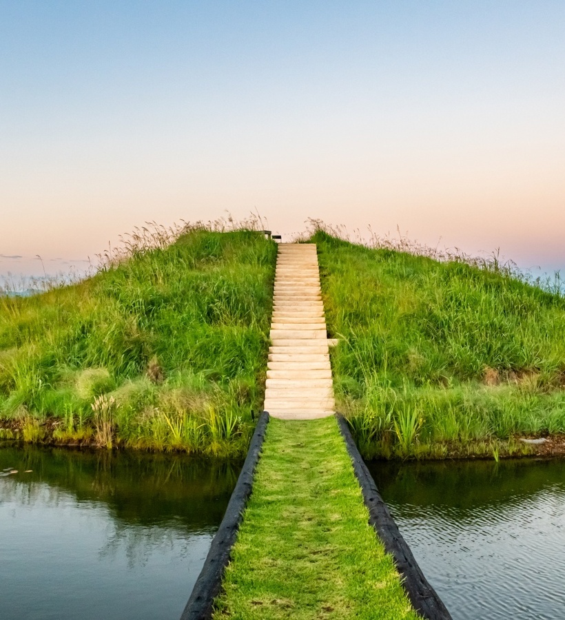 Grassy island reached by a narrow lawn bridge with stone steps, reflecting pond and pastel sunset - peaceful resort walk