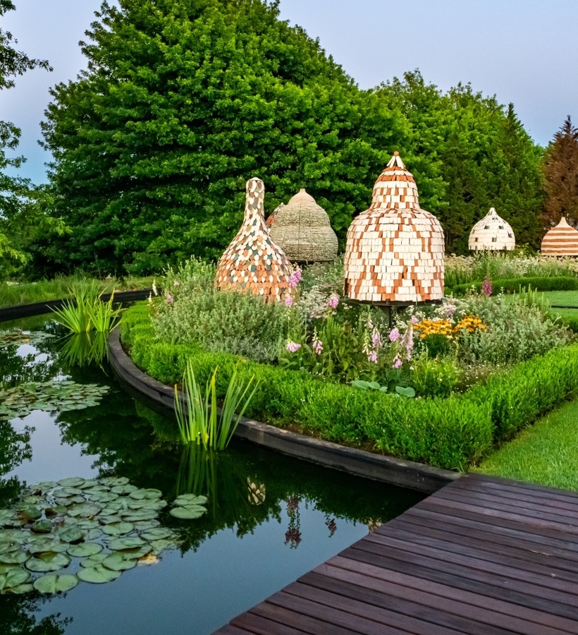 Manicured hotel garden with lily pond, wooden deck and woven beehive sculptures among trimmed boxwood and flowering beds.
