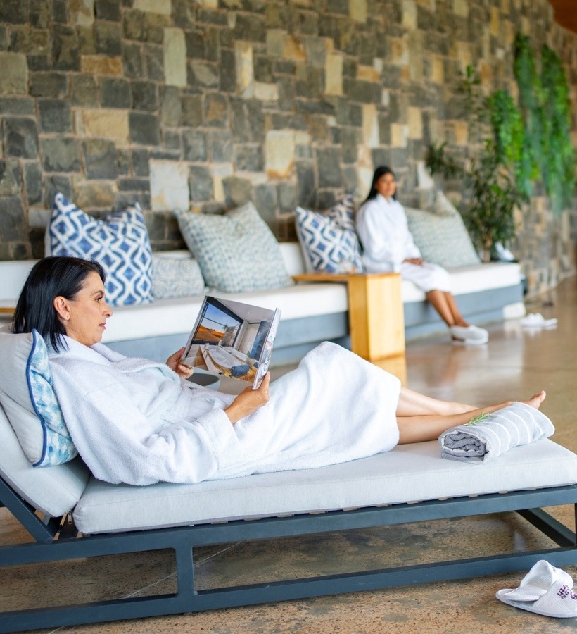Guest in white robe reading on a lounge beside an indoor pool, stone accent wall, cushioned seating and warm pendant lights.