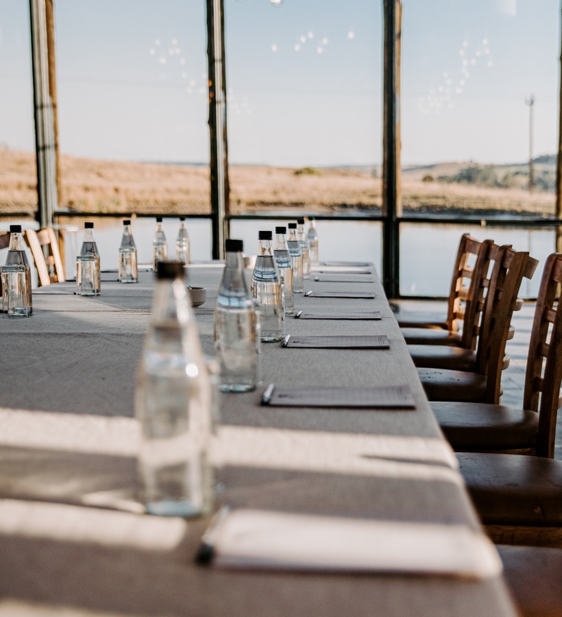 Linen meeting table with bottled water and notepads, wooden chairs by floor-to-ceiling windows overlooking golden fields.