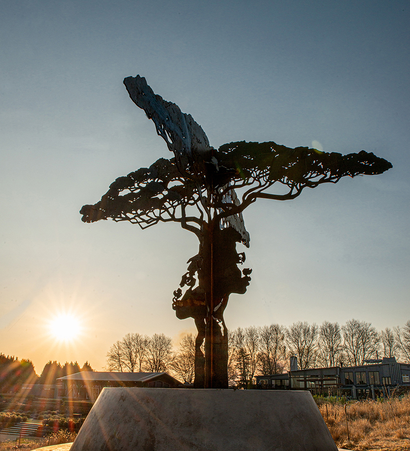 Metal tree sculpture with carved human profiles on a round concrete base, backlit by low sun over landscaped grounds and lodge.