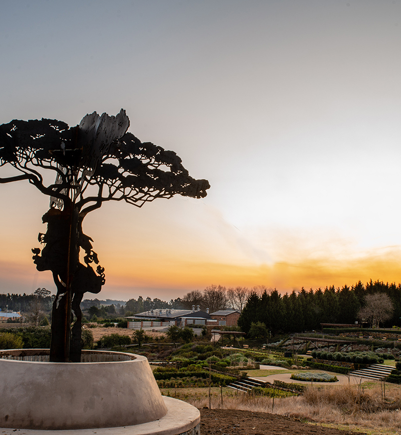 Silhouetted metal tree sculpture in a circular planter overlooking terraced hotel gardens, low guest buildings and a warm sunset sky.