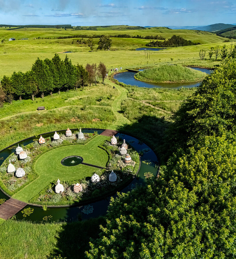 Circular ornamental garden with beehive sculptures and water moat, wooden bridges and paths across rolling green hills.