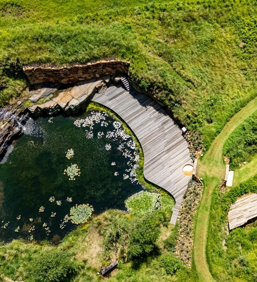 Curved wooden boardwalk beside a lily-pad pond with a small waterfall, grassy walking paths and a secluded pavilion
