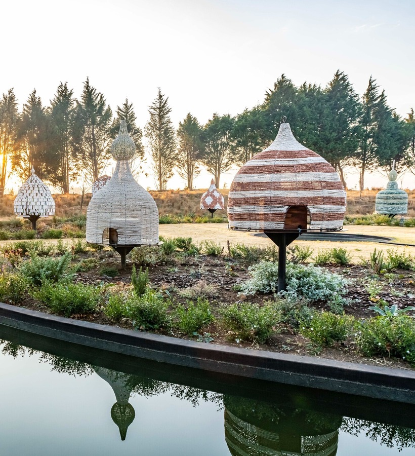 Serene hotel garden at sunrise with reed-woven dome huts on pedestals, reflecting pond, wooden deck and tree-lined horizon for peaceful relaxation