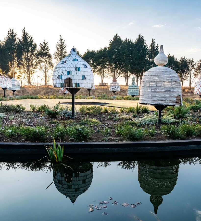 Woven beehive-style garden pavilions on pedestals, reflected in a calm pond at sunset with planted paths and pine trees.