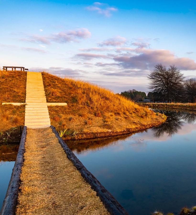 Stone steps up a grassy mound to a picnic bench, waterfront path and reflective pond at sunset