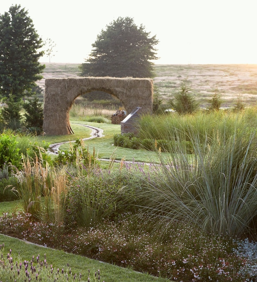 Sunlit hotel garden with winding grass path, lavender beds, a straw-arch feature and dunes on the horizon.