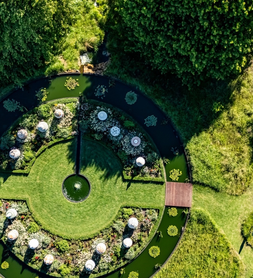 Aerial view of circular garden with central fountain, concentric flower beds, water moat and wooden bridges.