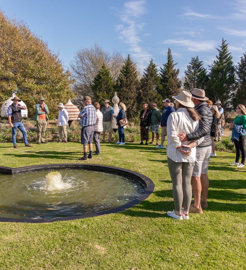 Guests on a sunny guided garden tour around a circular fountain amid sculptural outdoor art and mature trees.