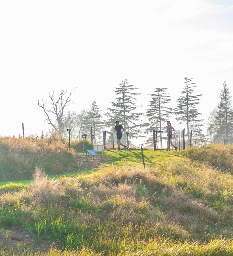 Sunlit hotel hillside with grassy nature trail and fencing; two guests jogging past a gated path amid tall trees, peaceful outdoor exercise setting.