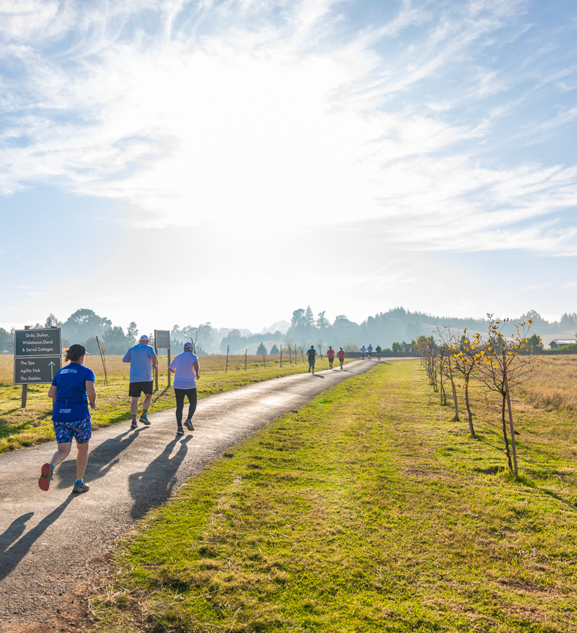 Guests jogging on sunlit hotel country path past young trees and grassy fields, with distant cottages and spa/stables sign.