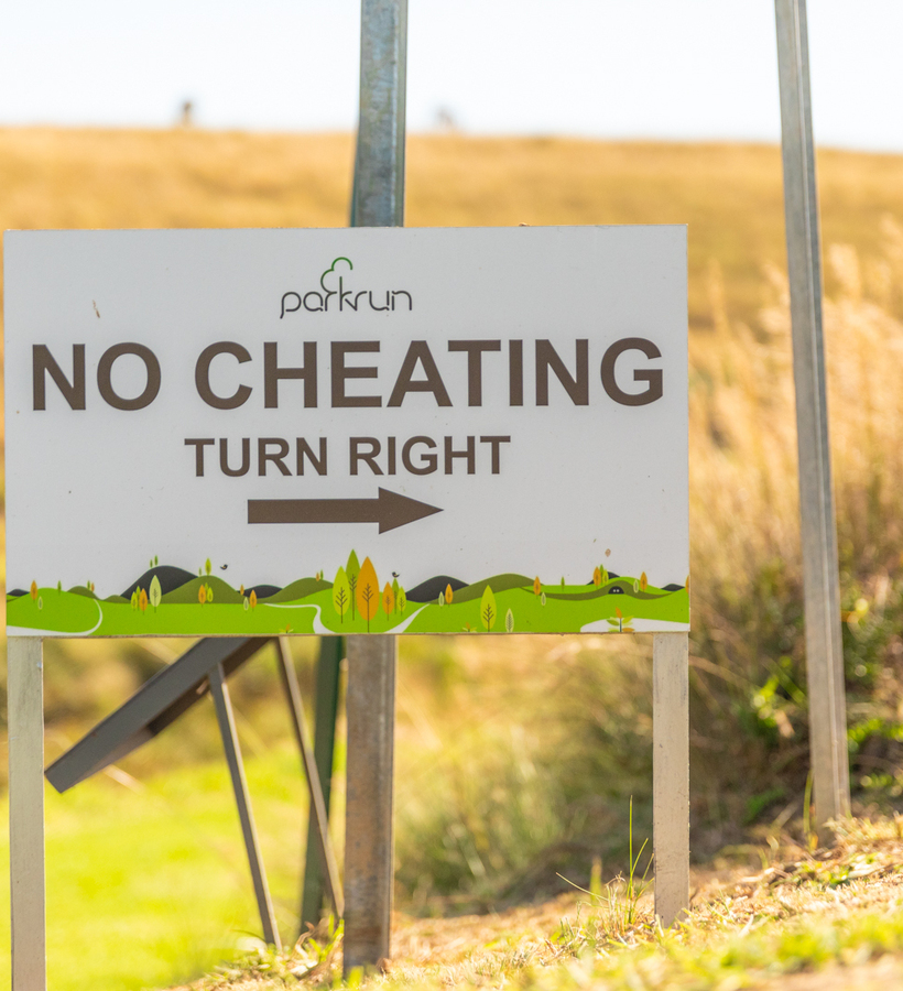 Parkrun sign: 'NO CHEATING TURN RIGHT' with arrow, mounted by a sunlit grassy trail beside dry, rolling hills
