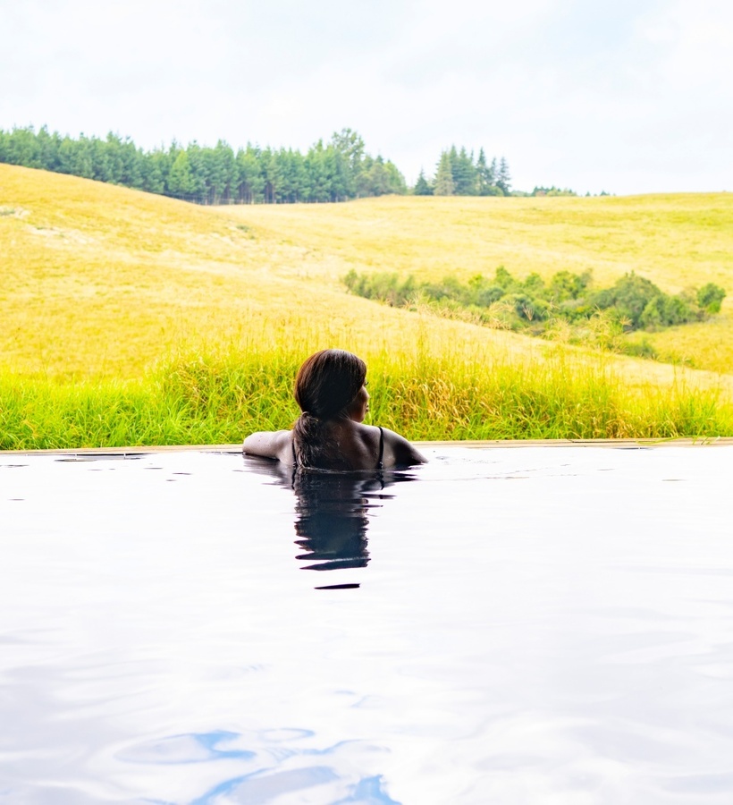 Guest relaxing in an indoor pool with open glass wall, overlooking golden rolling hills and a pine tree-lined ridge.