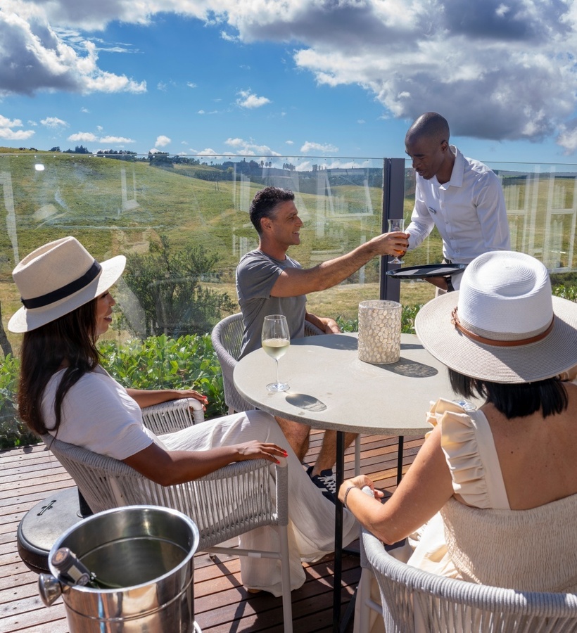 Rooftop terrace dining with waiter serving drinks to guests, wooden deck, glass railing and rolling green hillside views.