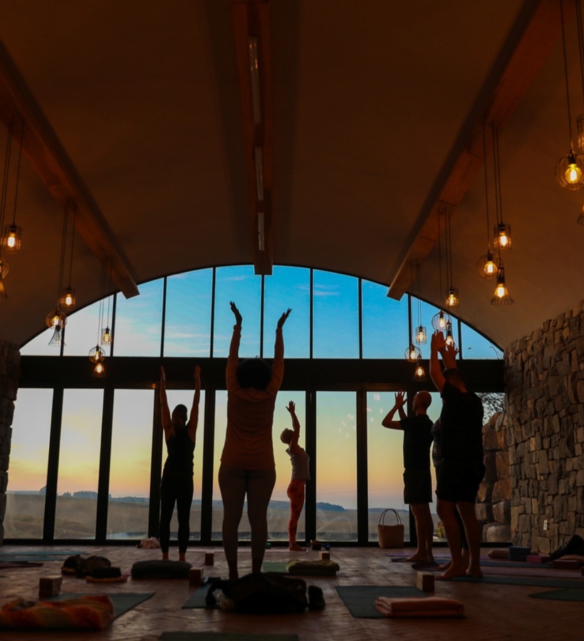 Sunset yoga class in a stone-walled wellness studio with arched glass wall, warm pendant lights and guests stretching arms.