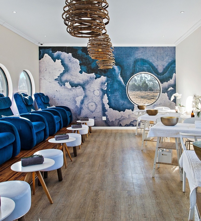 Bright spa lounge with plush blue recliners by round windows, manicure tables with brass bowls, orchids, wooden pendant lights and calming blue mural.