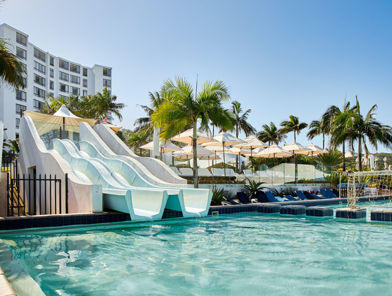 Resort pool with three white water slides feeding a sunlit swimming pool, lounge chairs under umbrellas, palm trees and hotel tower