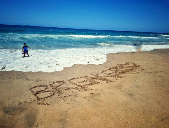 Beachfront with BREATHE written in sand, turquoise surf and a child wading at the shoreline under clear blue sky.
