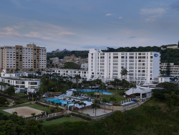 White multi-story coastal resort with balconies, palm grounds, pools, tented poolside restaurant and sand volleyball court.