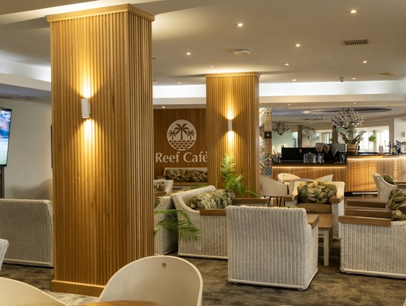 Reef Café lounge with wicker chairs, tropical cushions, warm wooden slatted columns, potted plants and illuminated bar.