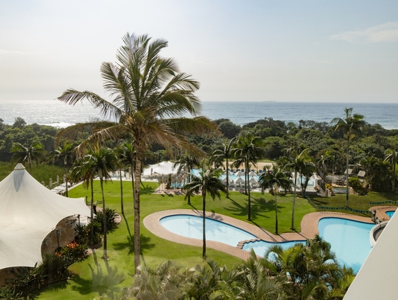Curving outdoor pools and palm‑dotted lawn with white event tent and poolside cabanas, beachfront view of ocean on the horizon.