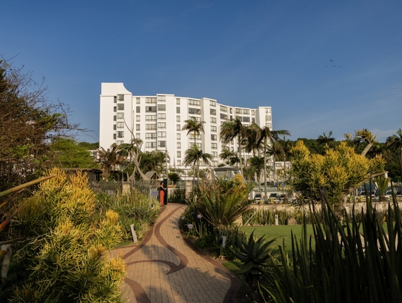 White beachfront Breakers Resort with palm-lined paved path through tropical gardens leading to pool and lounge chairs under blue sky.