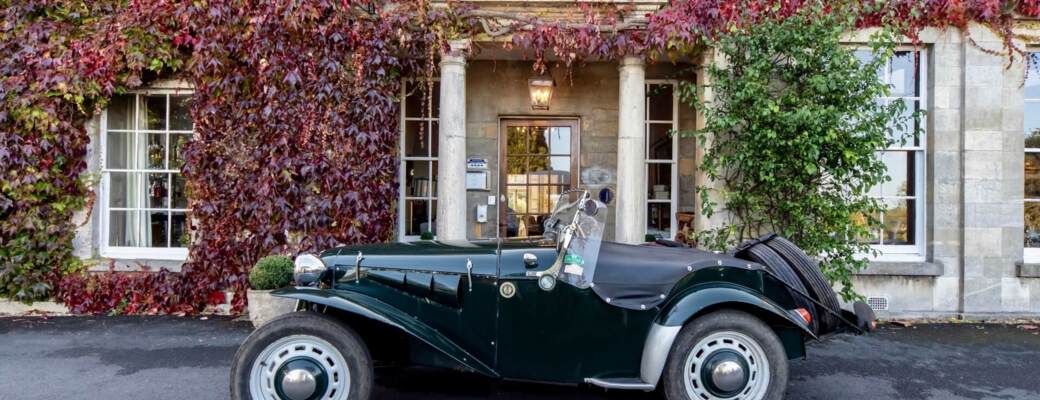 Classic vintage convertible parked in front of a historic stone hotel entrance covered in red ivy with tall columns and large windows