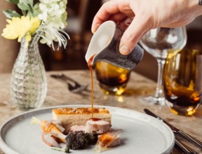 Hand pouring sauce over a plated gourmet pork dish with roasted vegetables in an elegant dining setting with glassware and fresh flowers