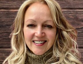 Smiling woman with shoulder-length blonde hair wearing a cozy brown turtleneck sweater against a wooden background