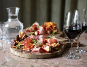 Charcuterie board with assorted cured meats, pickles, and garnishes paired with two glasses of red wine and a carafe of water on a marble table