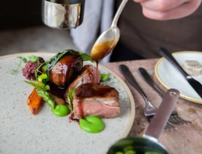 Sauce being drizzled over a plated gourmet dish with tender meat, asparagus, roasted carrot, and vibrant green purée