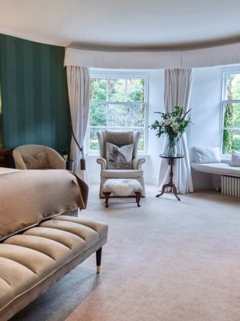 Spacious hotel bedroom with plush beige bedding, green striped walls, seating area by large windows, and elegant décor