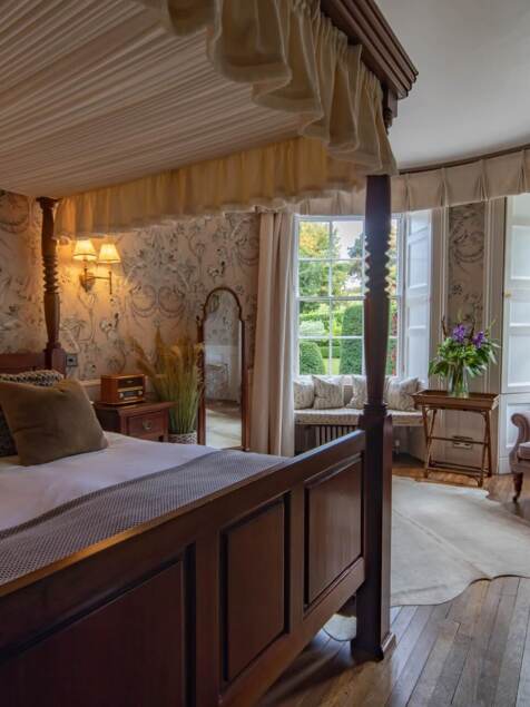 Cozy bedroom with four-poster bed, plush pillows, floral wallpaper, vintage radio, armchair, and garden views through large windows