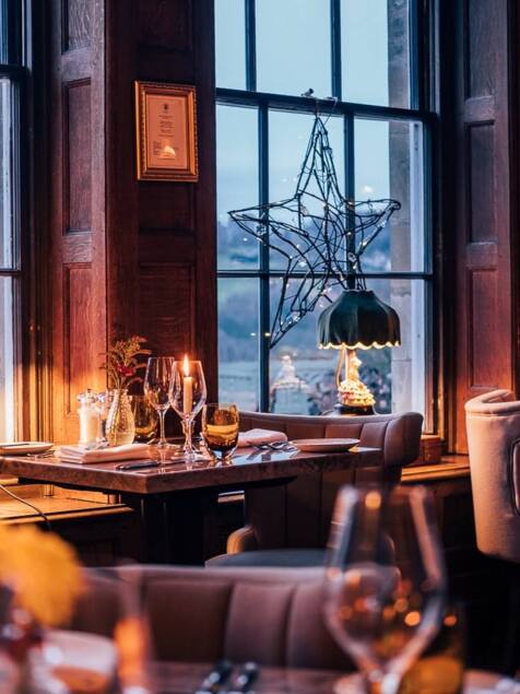 Cozy fine dining room with wood paneling, candlelit tables, plush chairs, decorative lamps, and large windows overlooking a countryside view