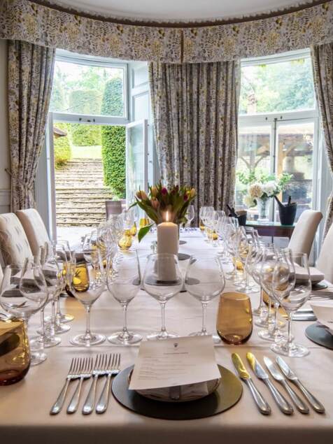 Elegant dining room with a long table set for a formal meal, floral centerpiece, candle, and patterned curtains by garden steps