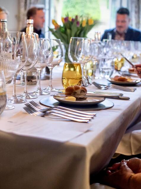 Elegant dining table set with multiple wine glasses, bread rolls, and butter, guests engaged in conversation in a cozy restaurant.