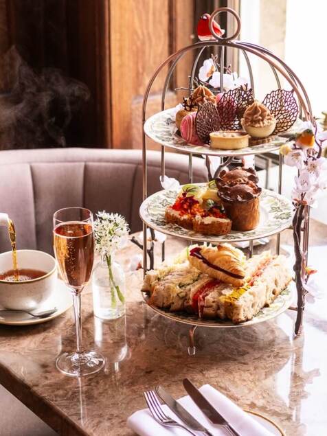 Afternoon tea setup with tiered platter of sandwiches, pastries, and desserts, steaming tea poured into a cup, and two glasses of sparkling wine