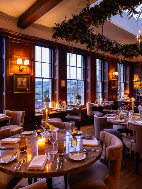 Cozy wood-paneled dining room with large windows, elegant round tables set for dinner, soft lighting, and festive greenery above