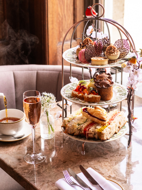 Afternoon tea setup with tiered tray of sandwiches, pastries, and sweets, accompanied by steaming tea and sparkling wine glasses