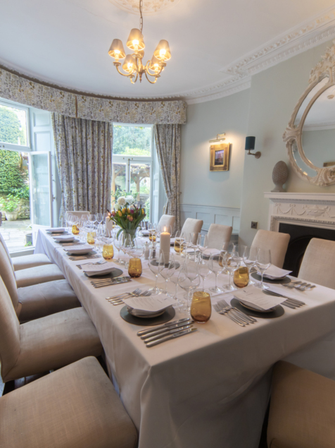 Elegant dining room with a long table set for a formal meal, featuring beige chairs, floral curtains, and ornate fireplace with decorative mirror