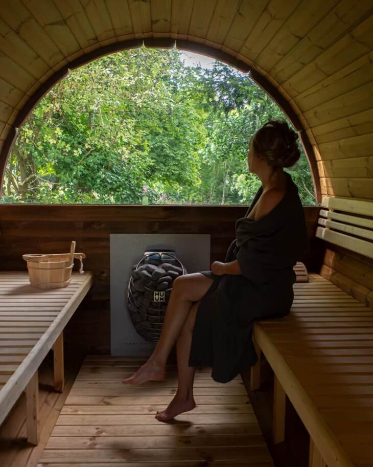 Guest wrapped in towel relaxing inside a wooden barrel sauna with large arched window overlooking lush green trees