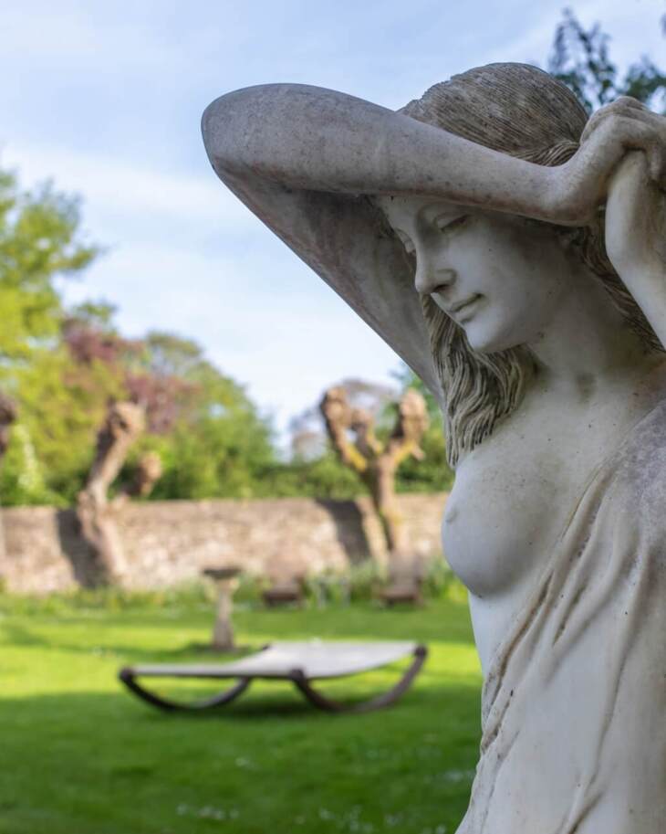 Elegant stone statue of a woman in a lush garden with trimmed trees and stone wall in the background
