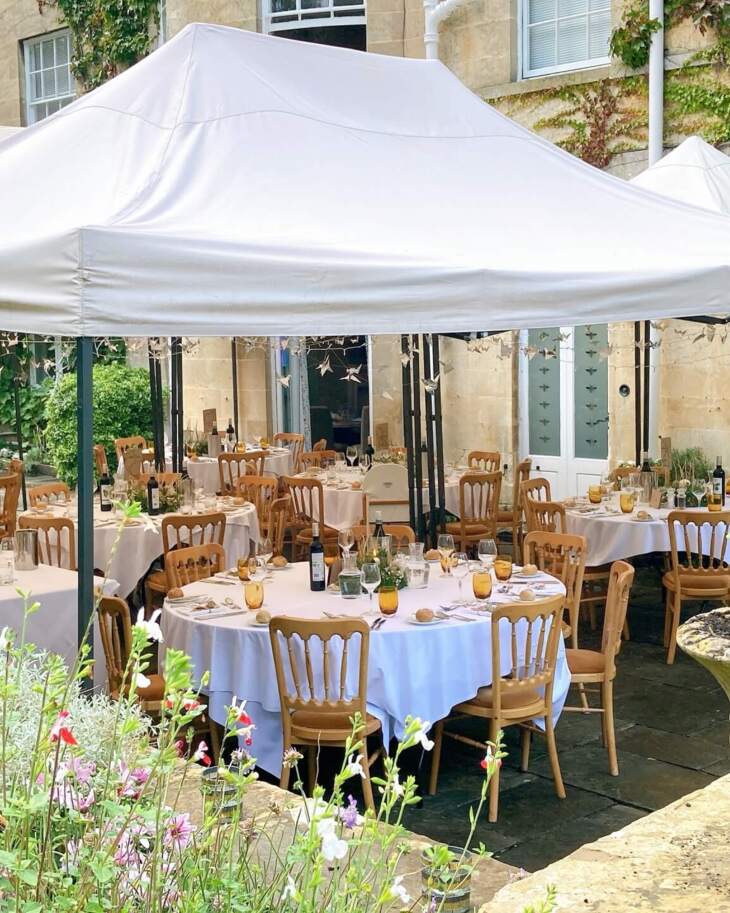 Outdoor hotel dining area under white canopy with round tables, wooden chairs, floral centerpieces, and warm glassware set for guests