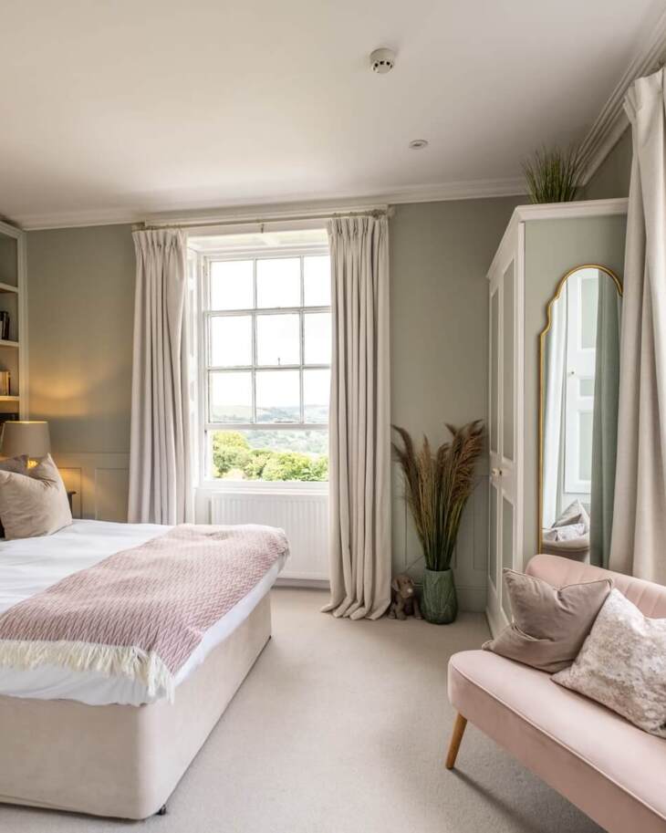 Bright hotel bedroom with large window, soft beige bed with multiple pillows, built-in shelves, pink loveseat, and elegant full-length mirror